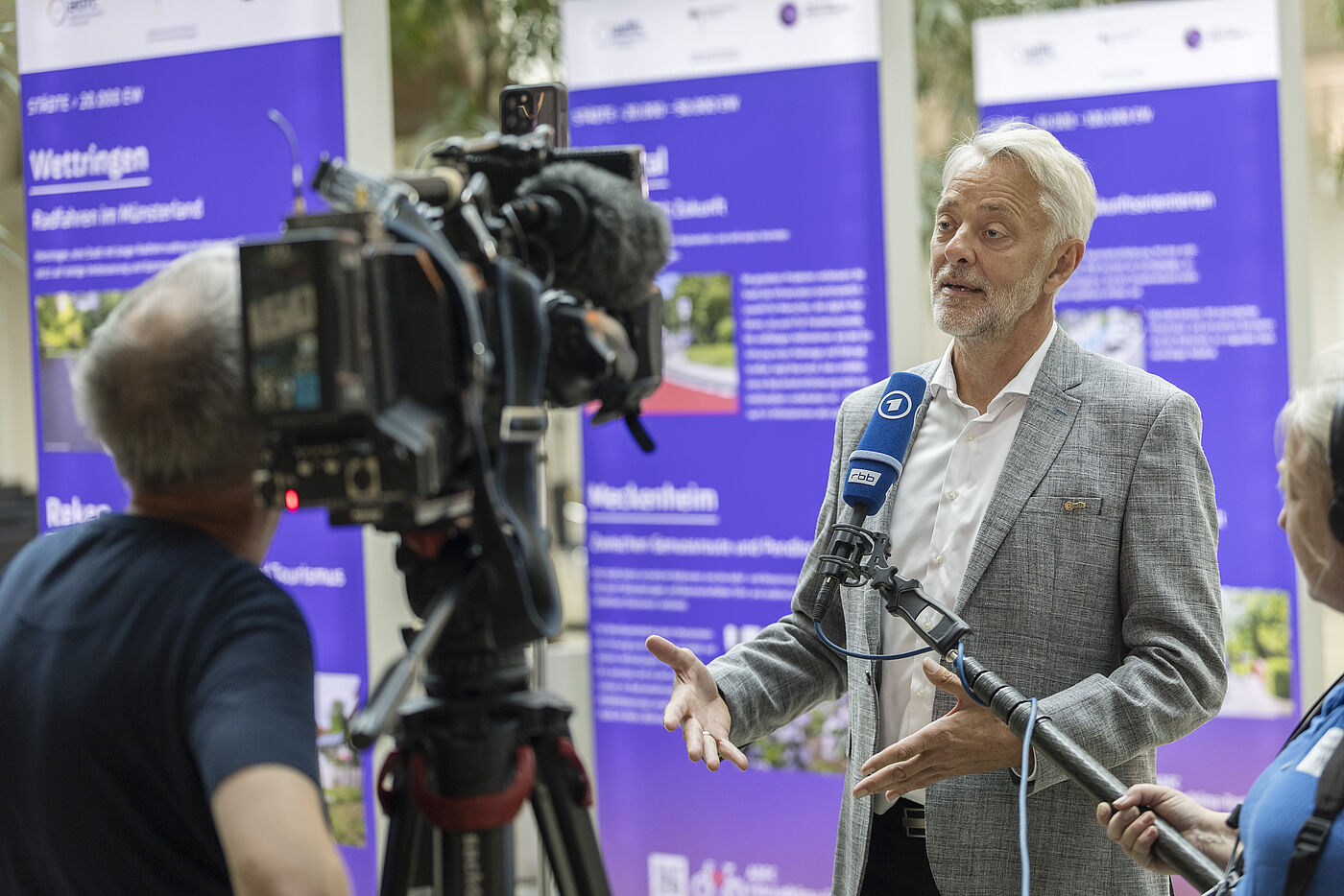 Frank Masurat, Mann mit grauen Haaren und Bart in hellgrauem Anzug, spricht in mehrere Mikrofone vor Fernsehkamera, im Hintergrund lila Banner mit Fahrradklima-Test-Informationen